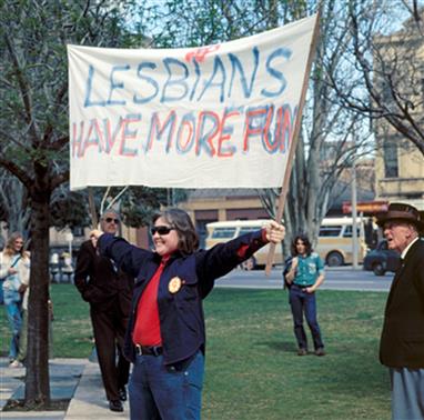 Woman in park holding banner reading Lesbians Have More Fun